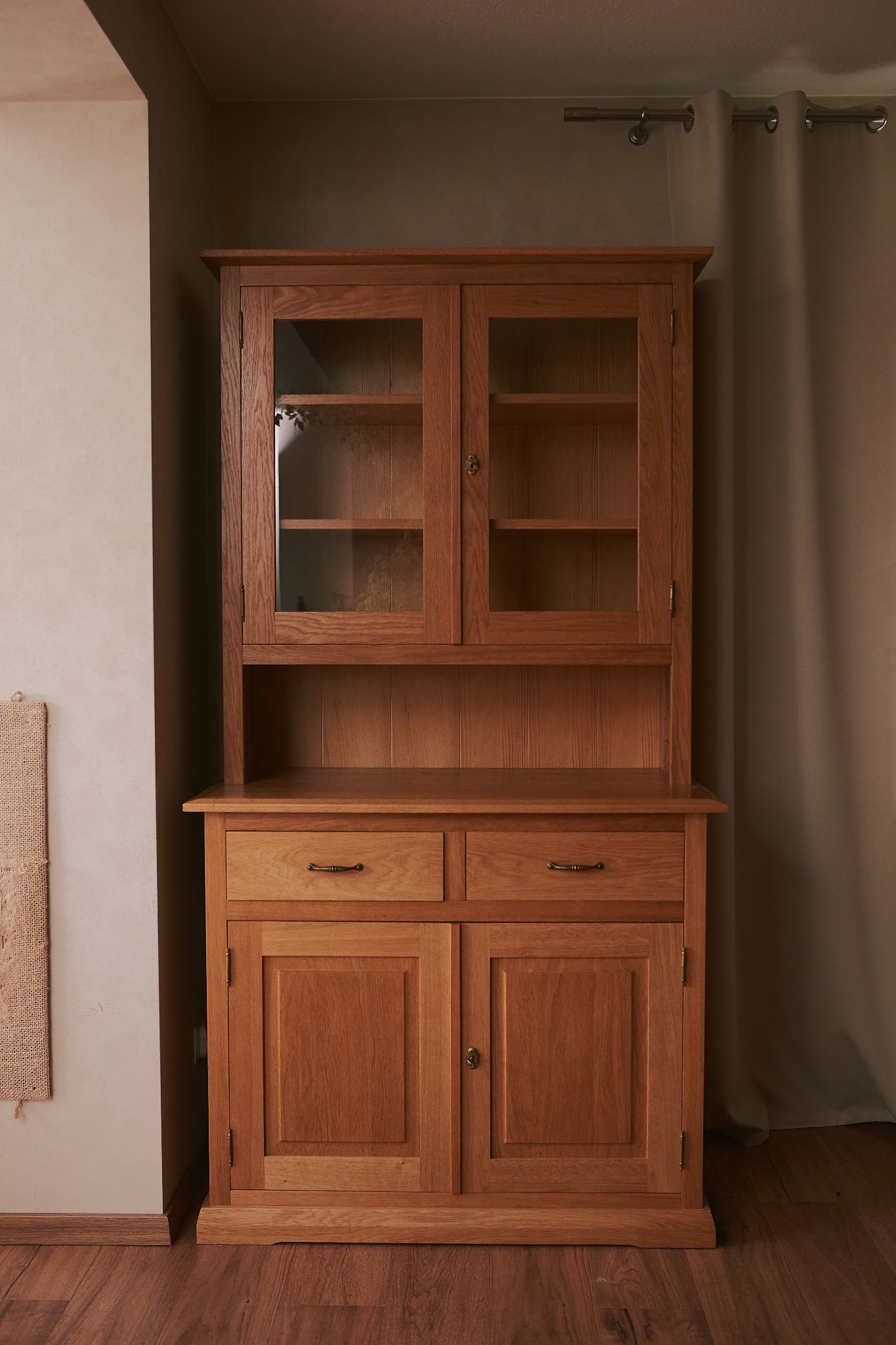 Drewniany kredens z przeszkloną nadstawką i szufladami, w ciepłej tonacji brązu, umieszczony we wnętrzu z zasłoną i drewnianą podłogą.