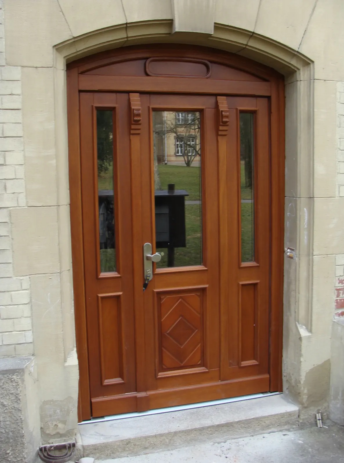 Eleganckie, drewniane drzwi wejściowe z przeszkleniami, osadzone w kamiennej futrynie. Widoczne detale architektoniczne i srebrna klamka z kluczem.