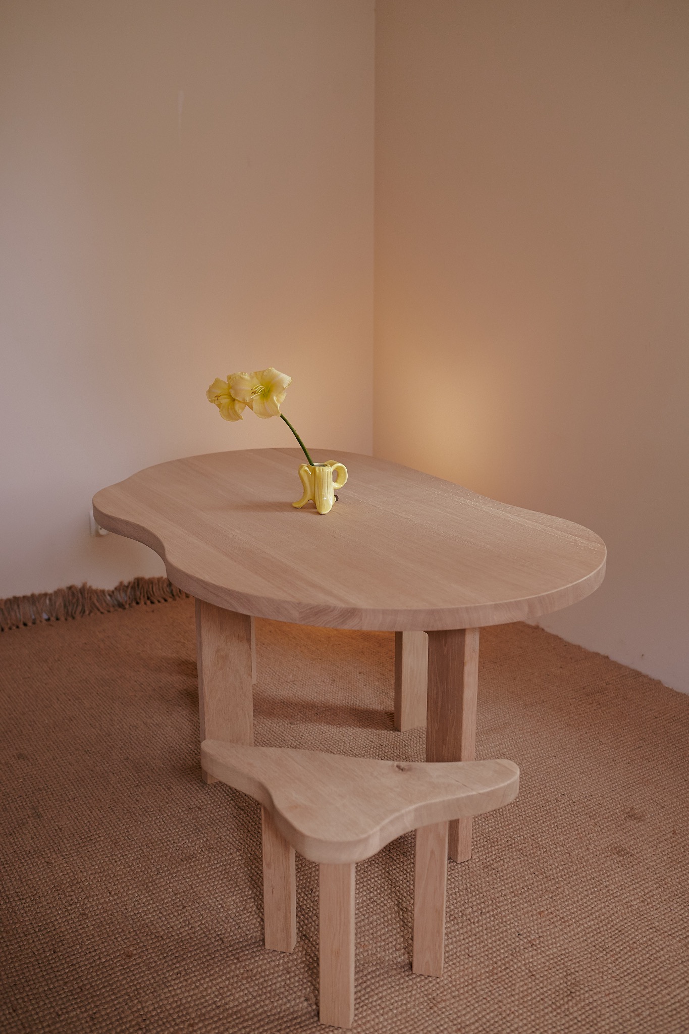 Drewniany stół o organicznym kształcie i stołek z jasnego drewna, na blacie żółty wazonik z kwiatami, całość na tle beżowej ściany i dywanu jutowego.