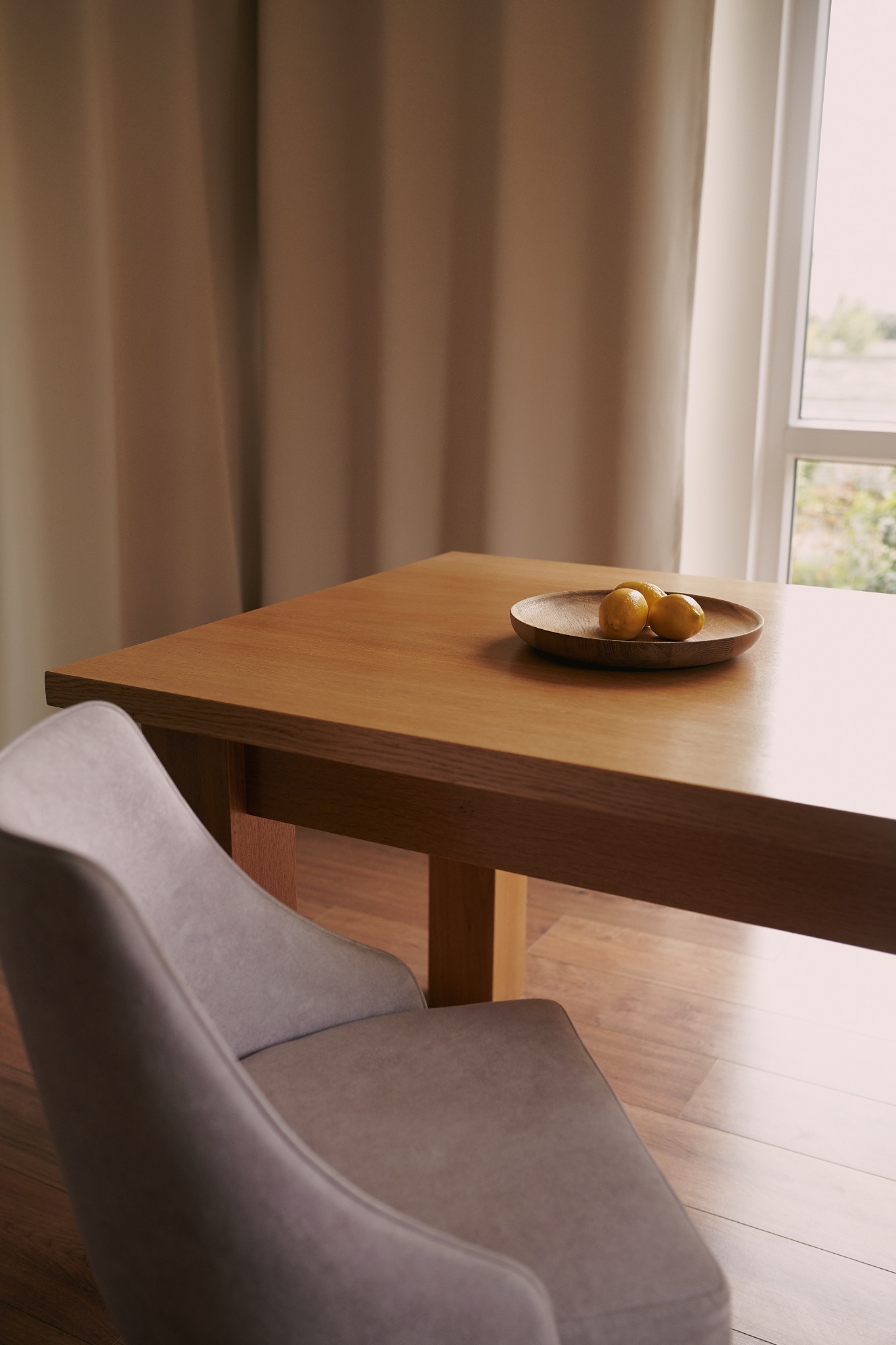 Drewniany stół z owocami na talerzu, obok krzesło z tapicerką w neutralnym odcieniu. W tle okno z zasłoną.