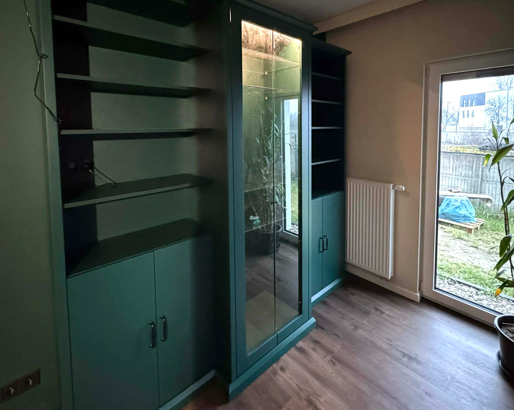 Zabudowa wnęki z szafkami i półkami w odcieniach zieleni, z przeszklonym segmentem pośrodku, na tle okna z widokiem na zewnątrz.