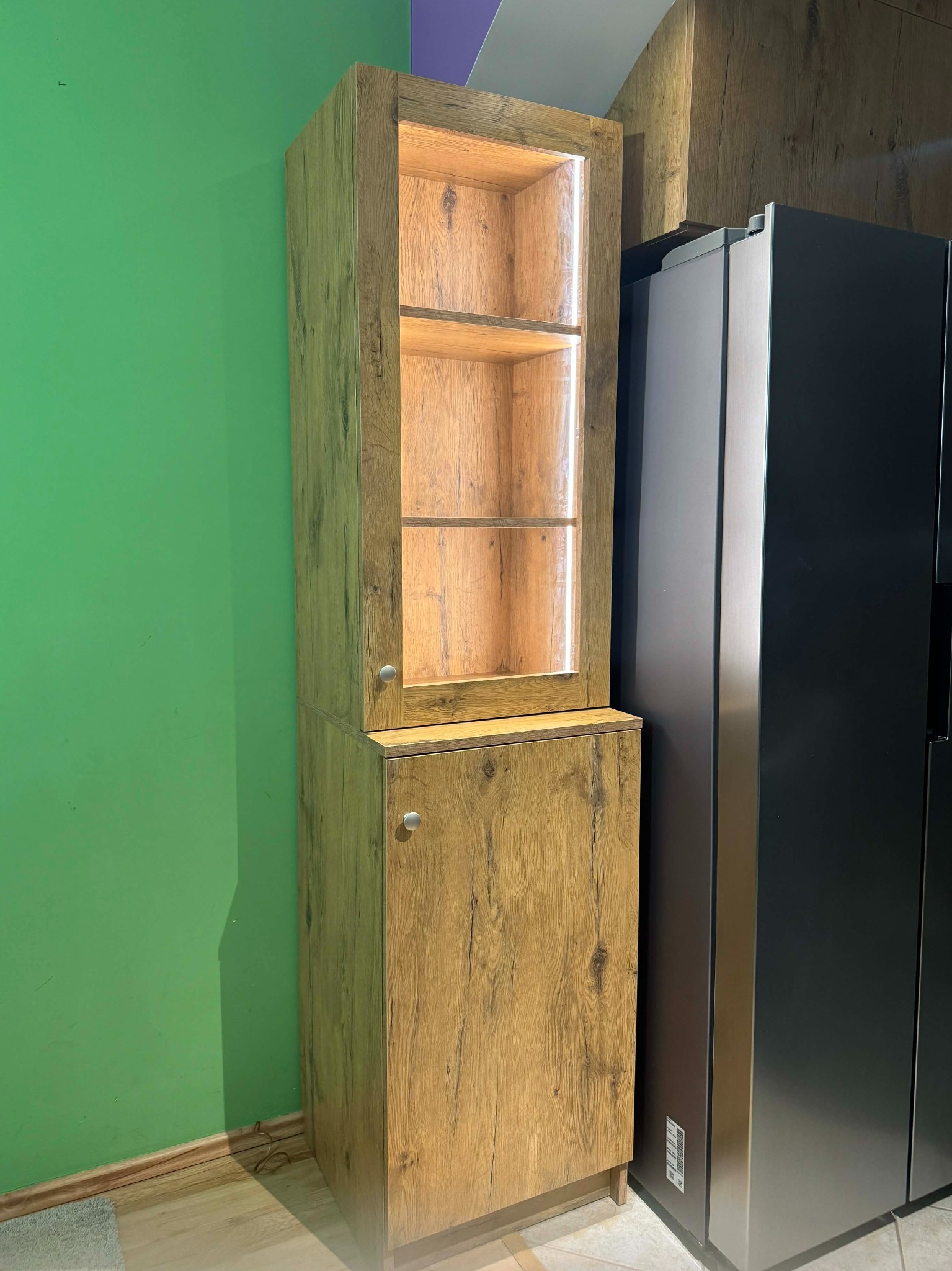 Wysoka, dwuczęściowa szafa z oświetleniem LED w górnej części, wykończona okleiną imitującą drewno, stoi przy zielonej ścianie obok czarnej lodówki.
