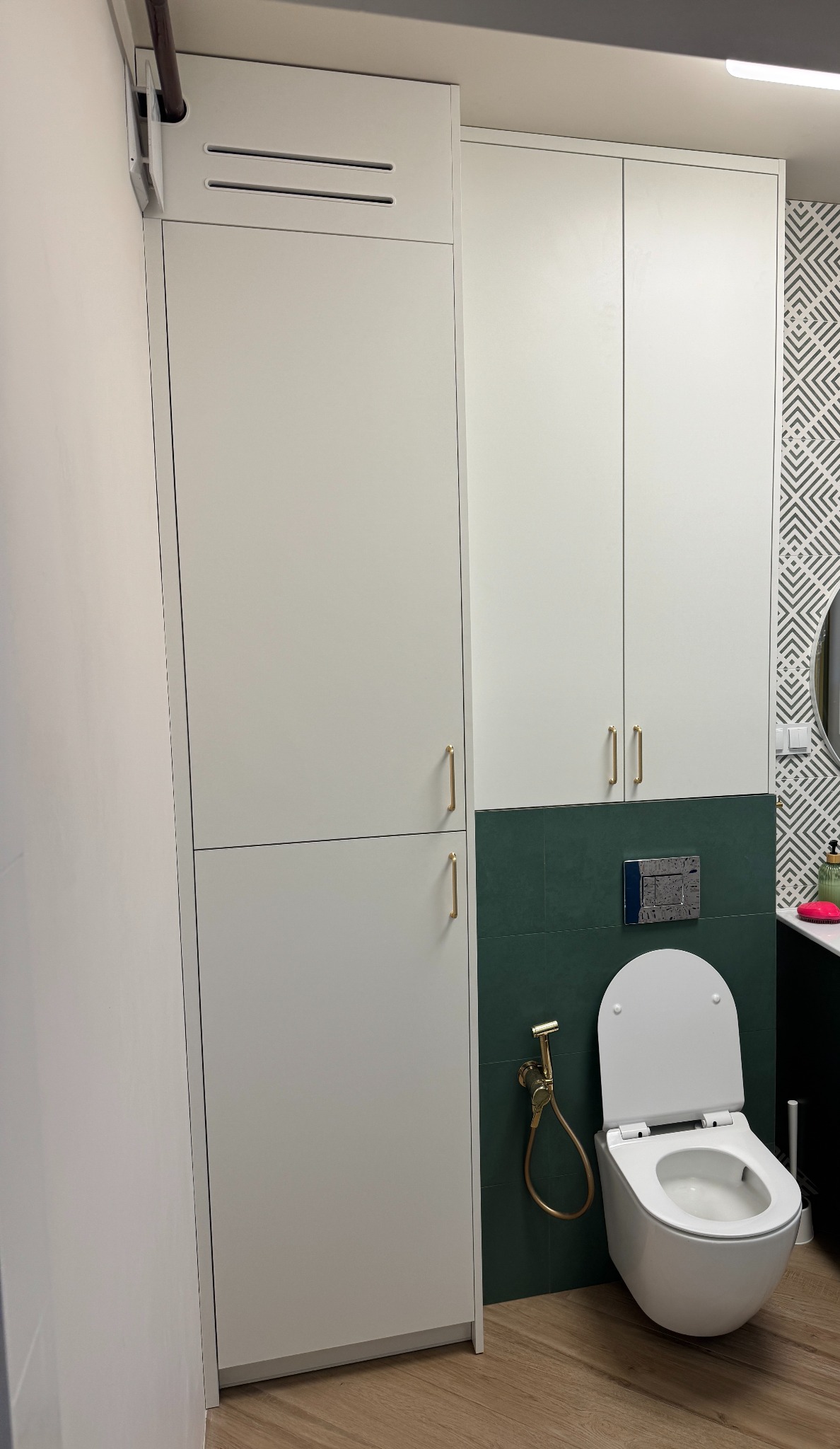 Biała szafa łazienkowa na wymiar z złotymi uchwytami, wisząca toaleta z bidetką na tle zielonej ściany i wzorzystej tapety. Nowoczesny design.