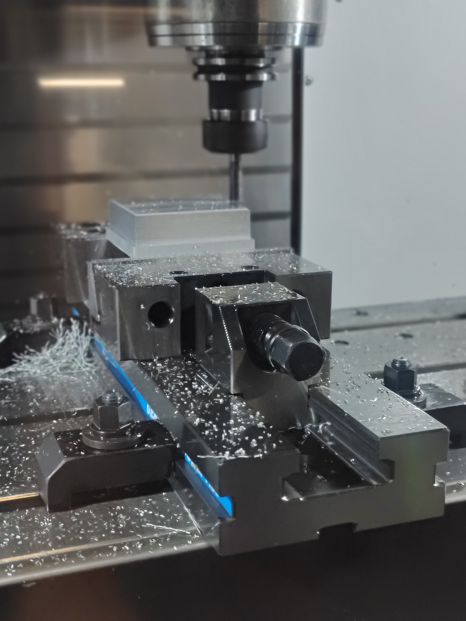 Obróbka CNC: Frezarka obrabia metalowy element w uchwycie, wióry na stole, oświetlenie halogenowe, precyzyjna obróbka skrawaniem.