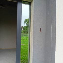 Grażyna Pilch Zakład Szklarski Szklane Chłopaki - Zamontowane okno przesuwne z widokiem na zewnątrz i surowym wykończeniem ścian. W tle zieleń trawnika i drzew.