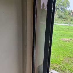 Grażyna Pilch Zakład Szklarski Szklane Chłopaki - Nowoczesne, przesuwne drzwi balkonowe z czarną ramą, widok na zielony ogród. Surowy stan wykończenia wnętrza, betonowe ściany. Paleta drewniana na podłodze.