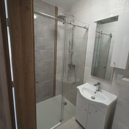 Grażyna Pilch Zakład Szklarski Szklane Chłopaki - Nowoczesna kabina prysznicowa ze szkła hartowanego w łazience wykończonej w szaro-białej kolorystyce z drewnianymi akcentami drzwi.