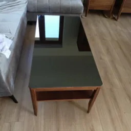 Szklany stolik kawowy z drewnianą ramą, ustawiony w salonie obok szarej sofy i drewnianej komody, odbijający w blacie okno i fragment sufitu.