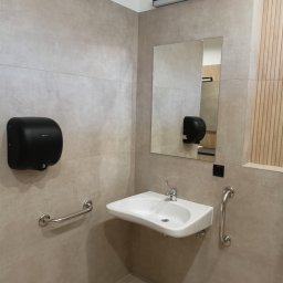 Grażyna Pilch Zakład Szklarski Szklane Chłopaki - Nowoczesna łazienka publiczna z umywalką, lustrem i uchwytami dla niepełnosprawnych. Beżowe płytki i dyskretne oświetlenie tworzą minimalistyczny design.