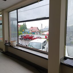 Grażyna Pilch Zakład Szklarski Szklane Chłopaki - Montaż okna: widok z wnętrza na ulicę przez surowe ramy okienne. Narzędzia i plany na parapecie, sylwetka osoby po lewej. Etap prac szklarskich.