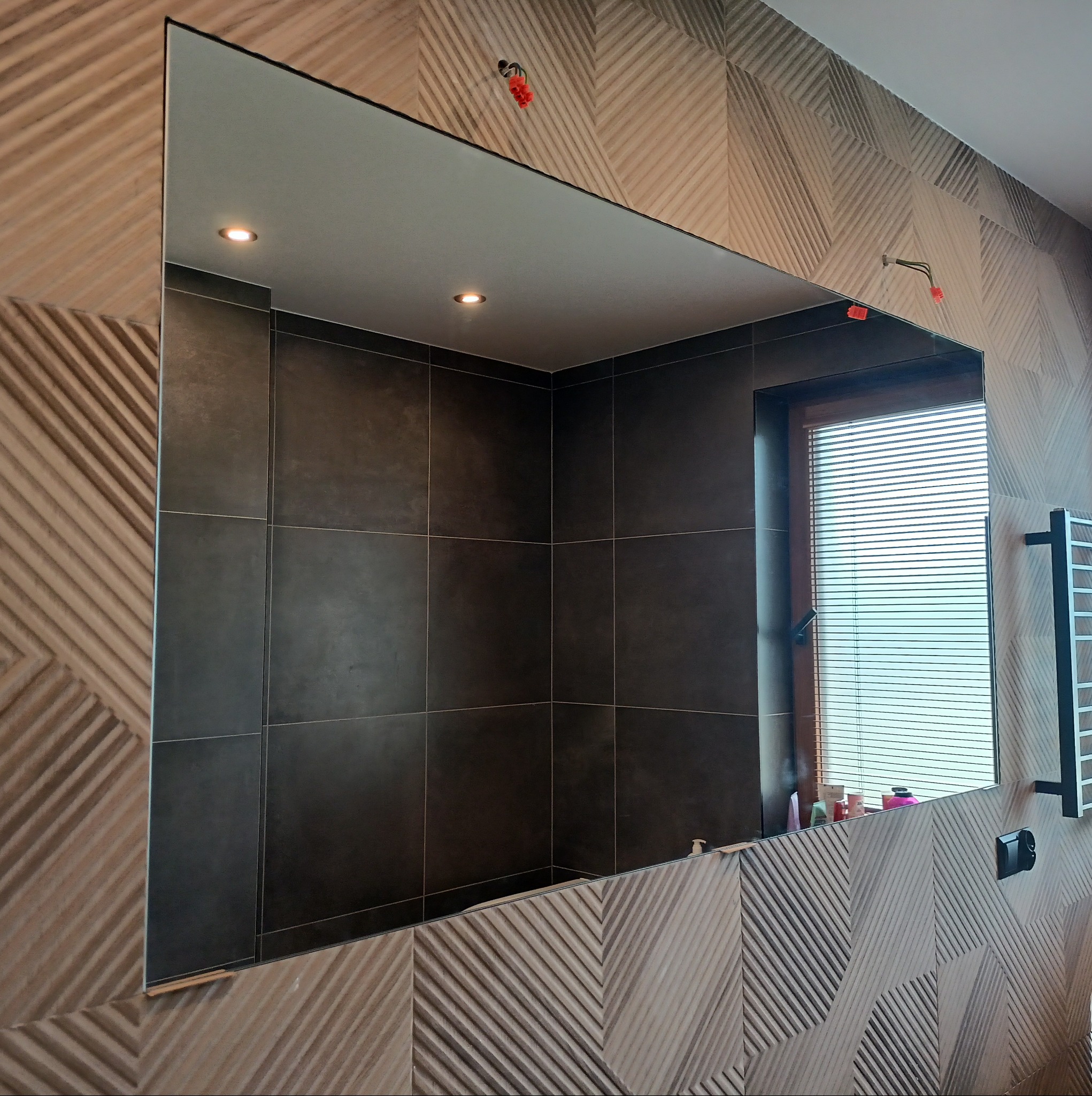 Prostokątne lustro w łazience z dekoracyjną ścianą, odbijające wnętrze z oknem w rogu. Widoczne elementy instalacji elektrycznej i grzejnik.