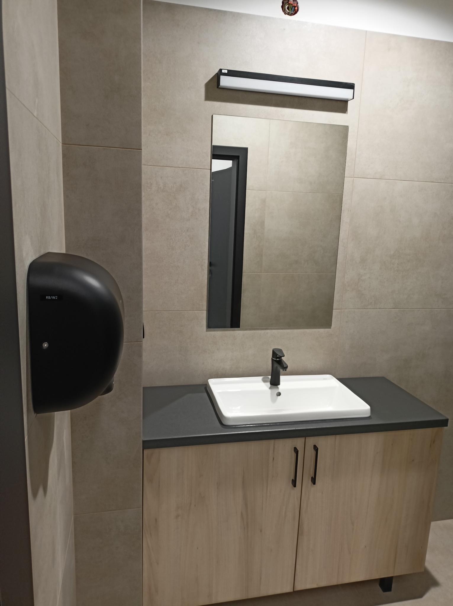 Nowoczesna łazienka z beżowymi płytkami, lustrem i białą umywalką na szafce z drewnopodobnymi frontami. Czarny dozownik do rąk po lewej stronie.