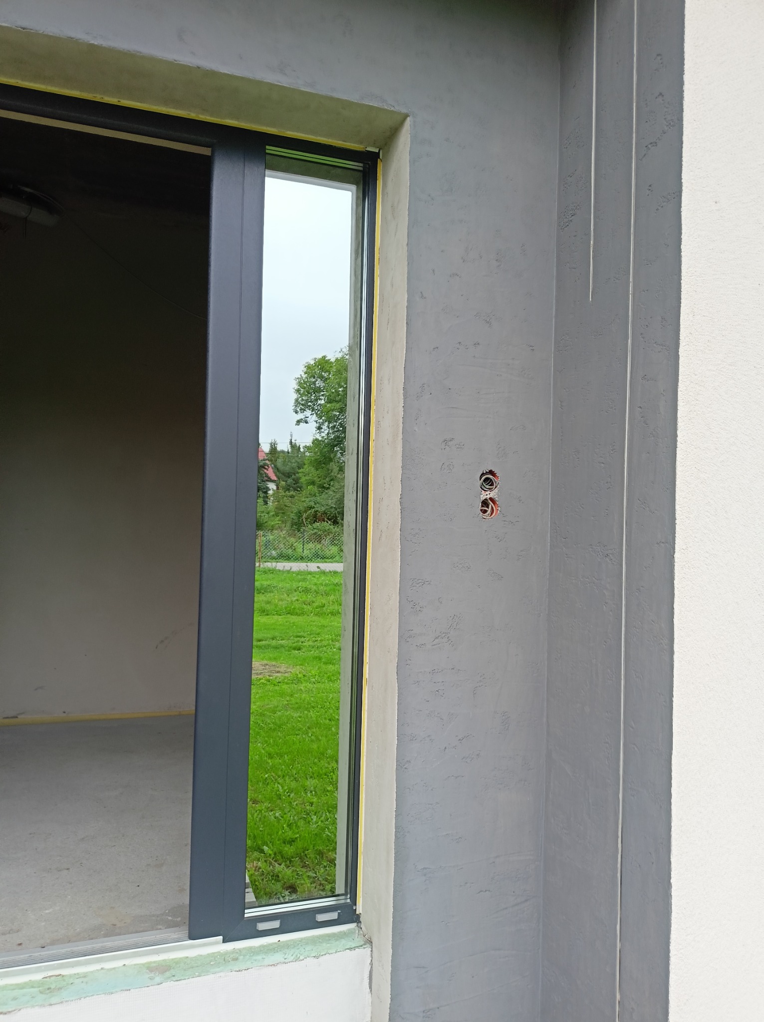 Zamontowane okno przesuwne z widokiem na zewnątrz i surowym wykończeniem ścian. W tle zieleń trawnika i drzew.