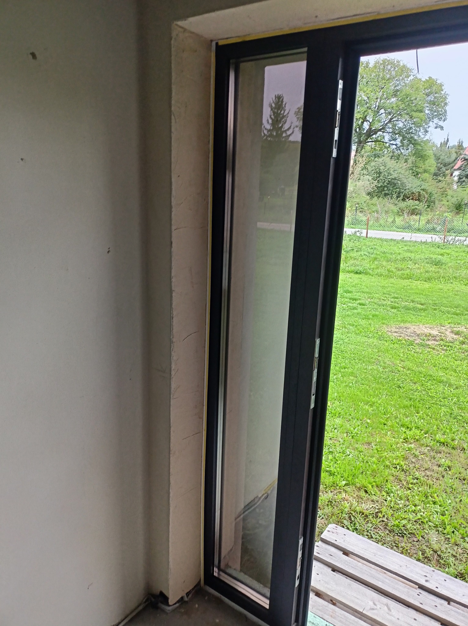 Nowoczesne, przesuwne drzwi balkonowe z czarną ramą, widok na zielony ogród. Surowy stan wykończenia wnętrza, betonowe ściany. Paleta drewniana na podłodze.