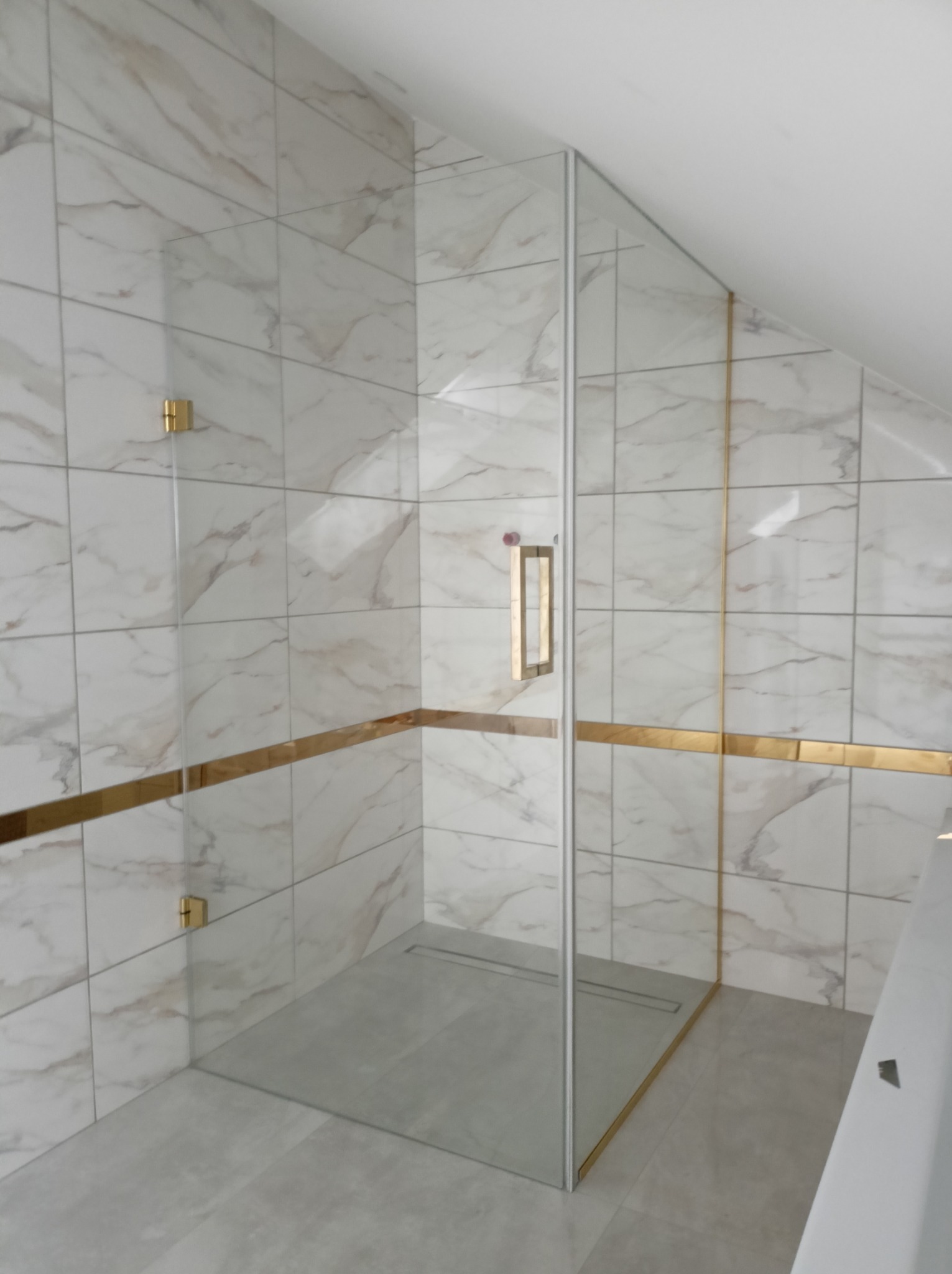 Kabina prysznicowa ze szklanymi drzwiami, złote okucia i listwa dekoracyjna na tle marmurowych płytek w łazience na poddaszu.