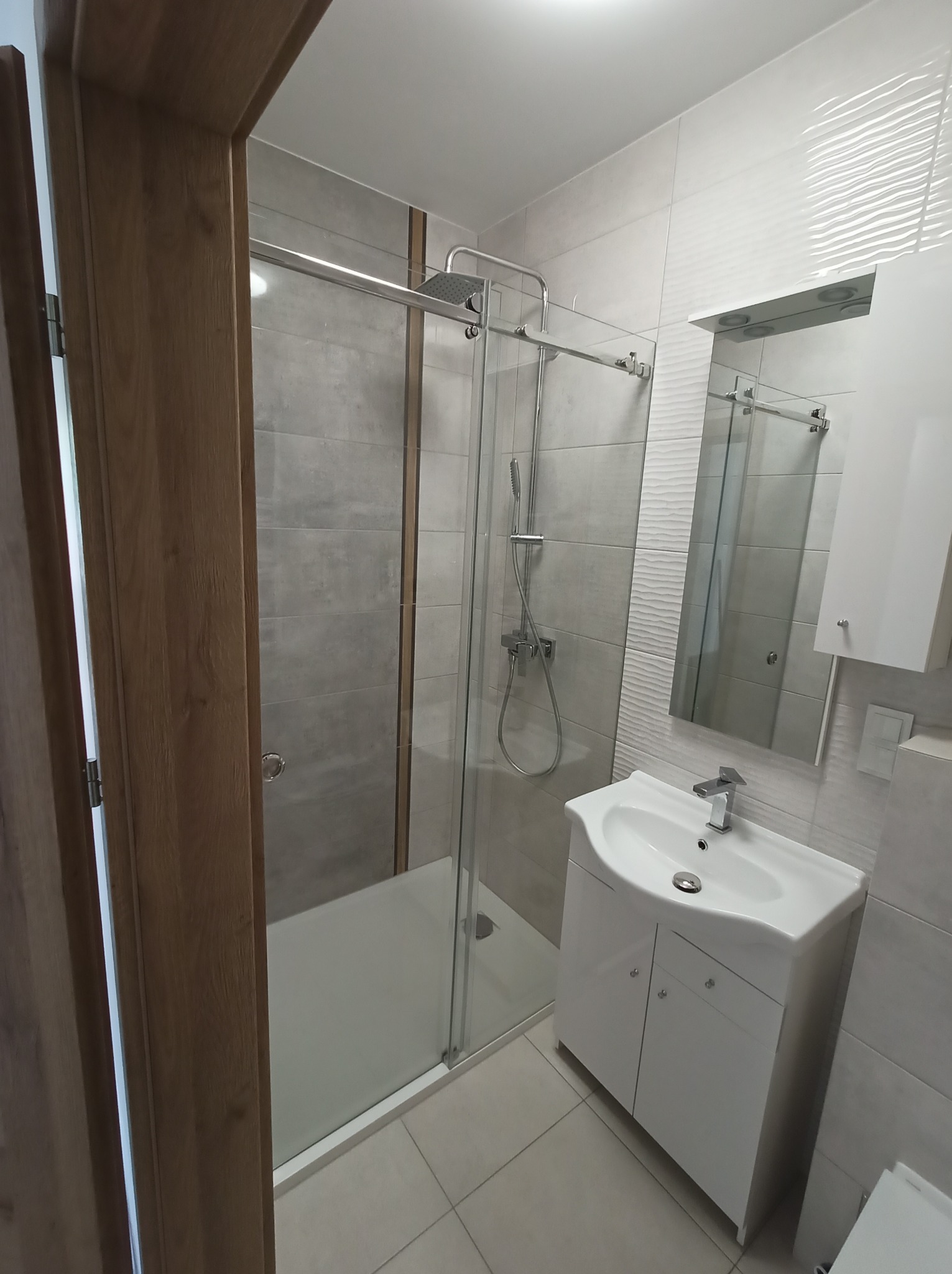 Nowoczesna kabina prysznicowa ze szkła hartowanego w łazience wykończonej w szaro-białej kolorystyce z drewnianymi akcentami drzwi.