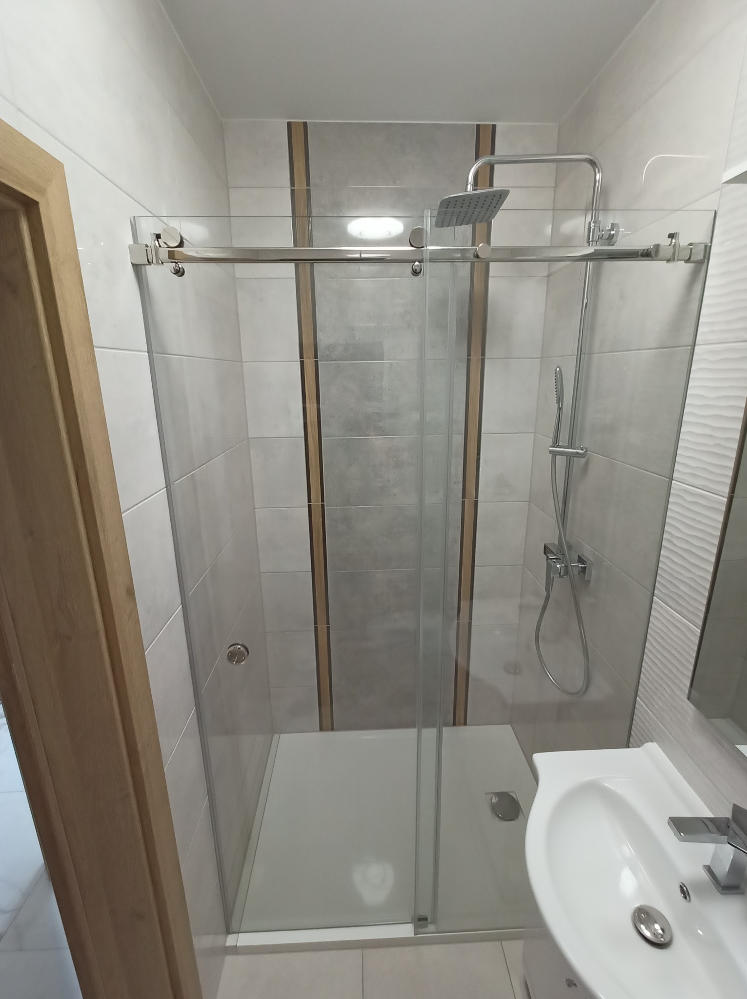 Nowoczesna kabina prysznicowa ze szklanymi drzwiami przesuwnymi, chromowane okucia, biały brodzik i umywalka. Widok z boku, neutralne oświetlenie.