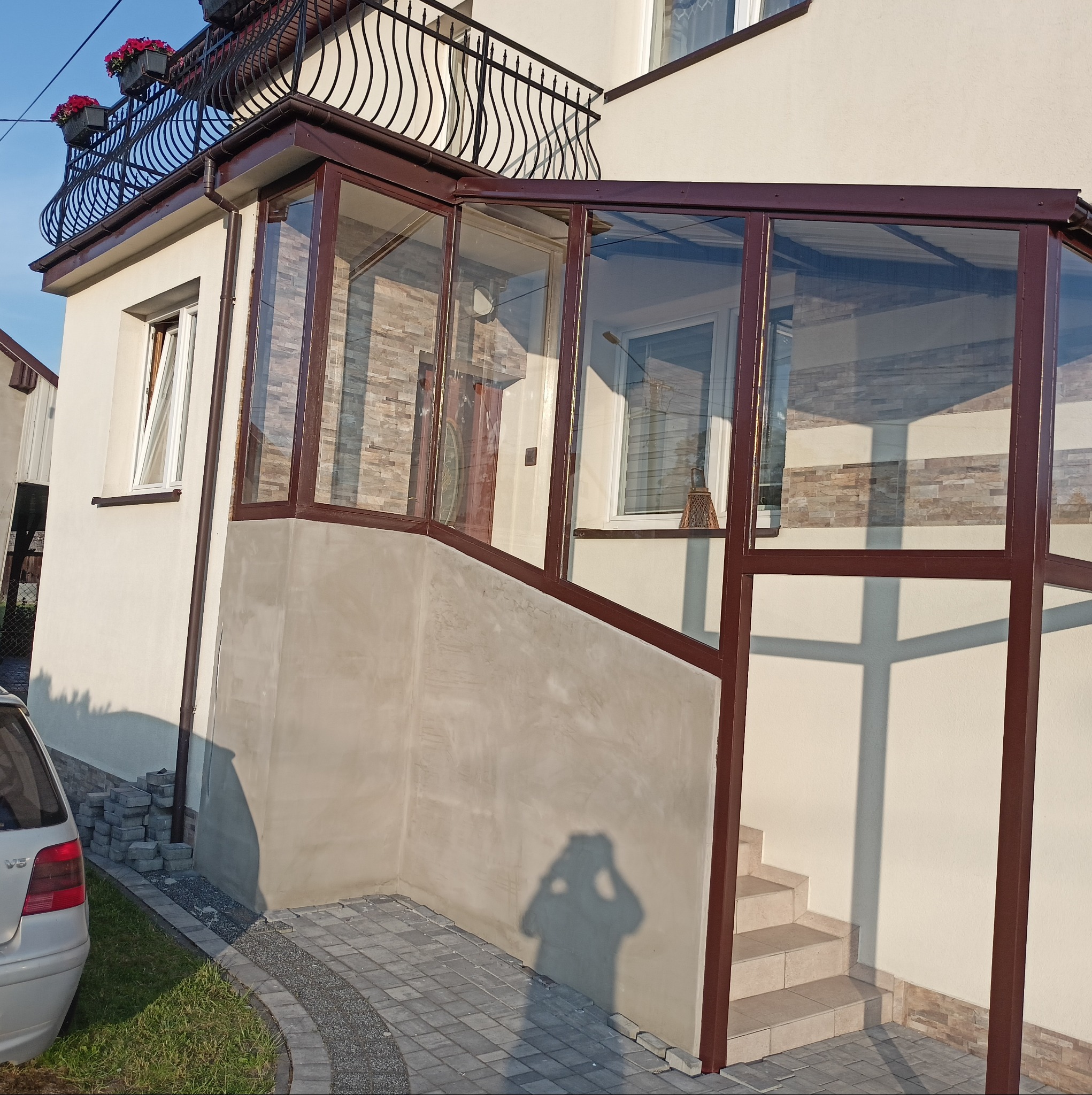 Zabudowa wejścia do domu z brązową ramą i przeszkleniami. Widoczne schody wejściowe i fragment elewacji. Na górze balkon z ozdobną, metalową balustradą.