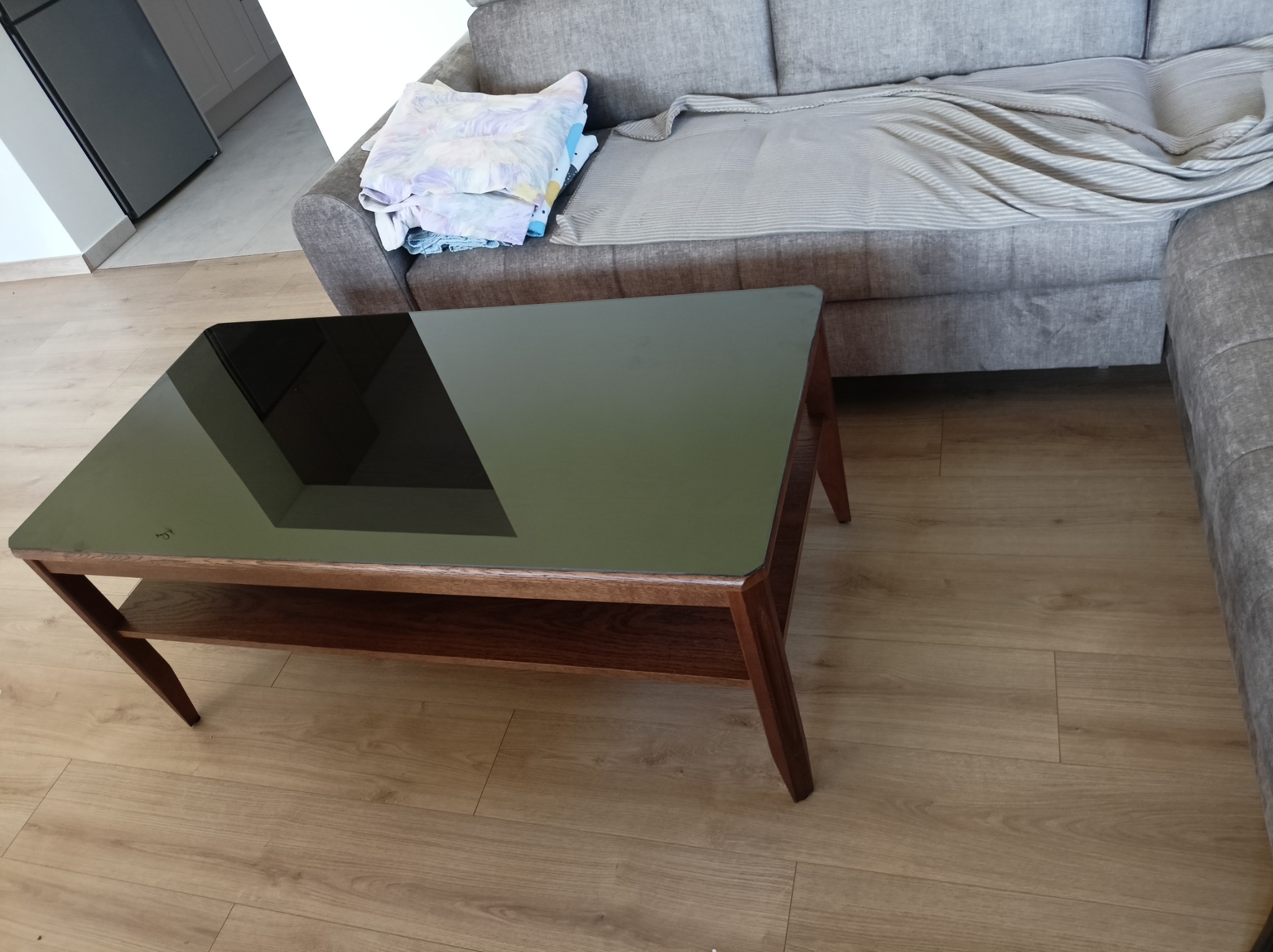 Stolik kawowy z brązowymi nogami i czarnym, szklanym blatem. Obok kanapa z narzutą. Widok z perspektywy osoby stojącej. Parkiet na podłodze.