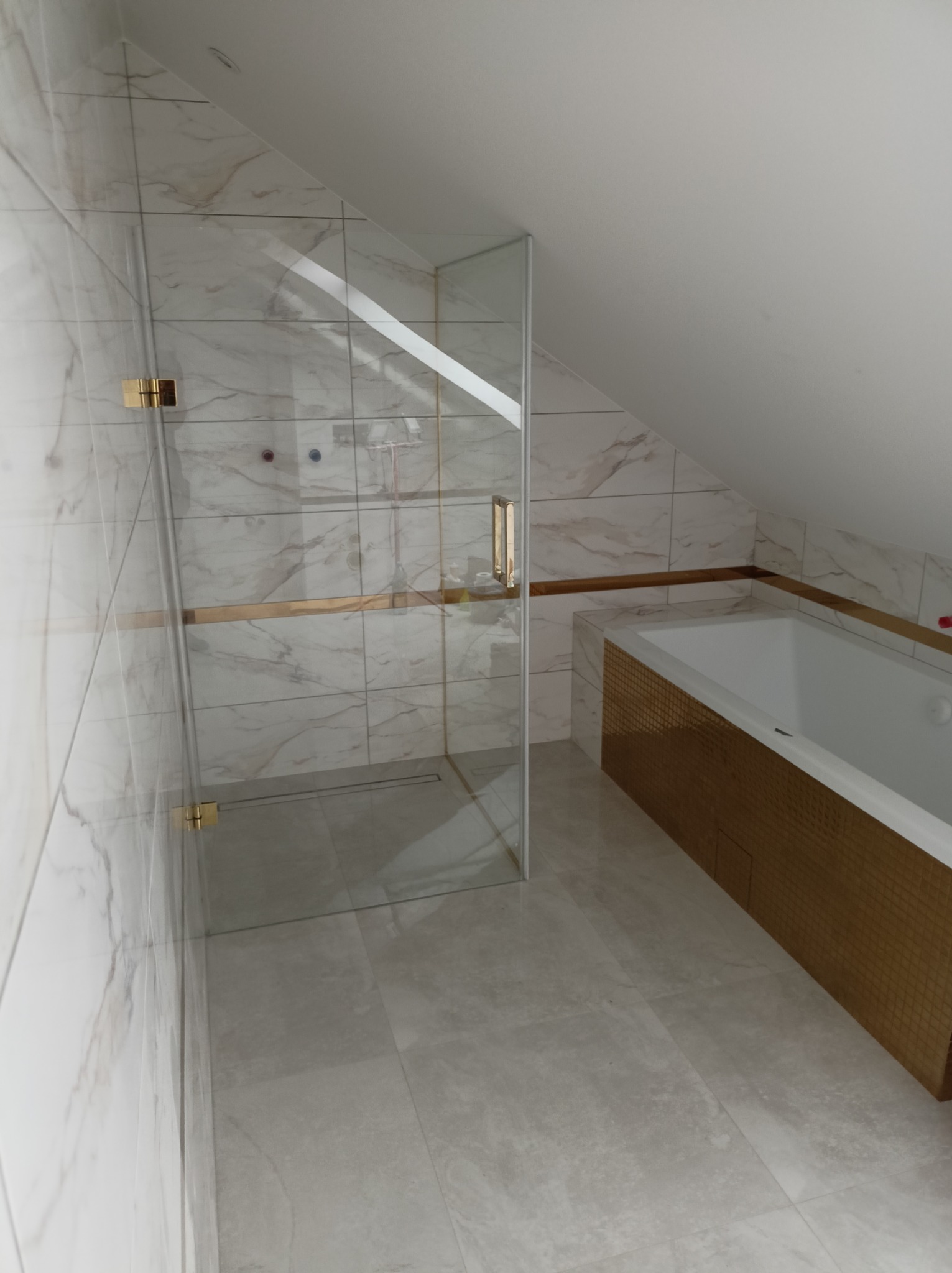 Kabina prysznicowa ze złotymi akcentami w łazience na poddaszu, wykończonej marmurowymi płytkami i mozaiką. Widoczny fragment wanny.