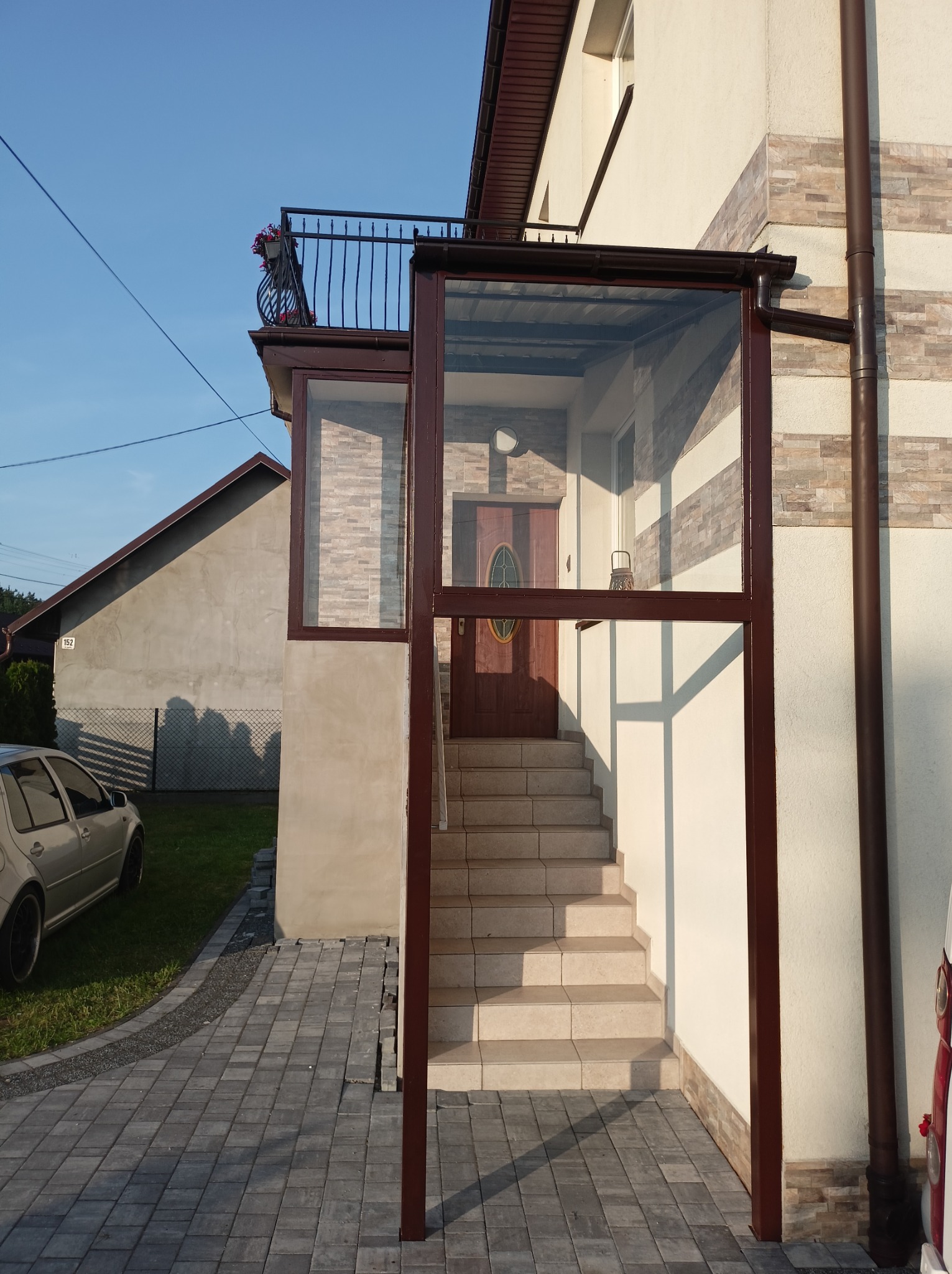 Szklana zabudowa wejścia do domu z brązowymi ramami, schody z jasnych płytek, elewacja w odcieniach beżu i szarości. Widoczny fragment balustrady balkonowej.