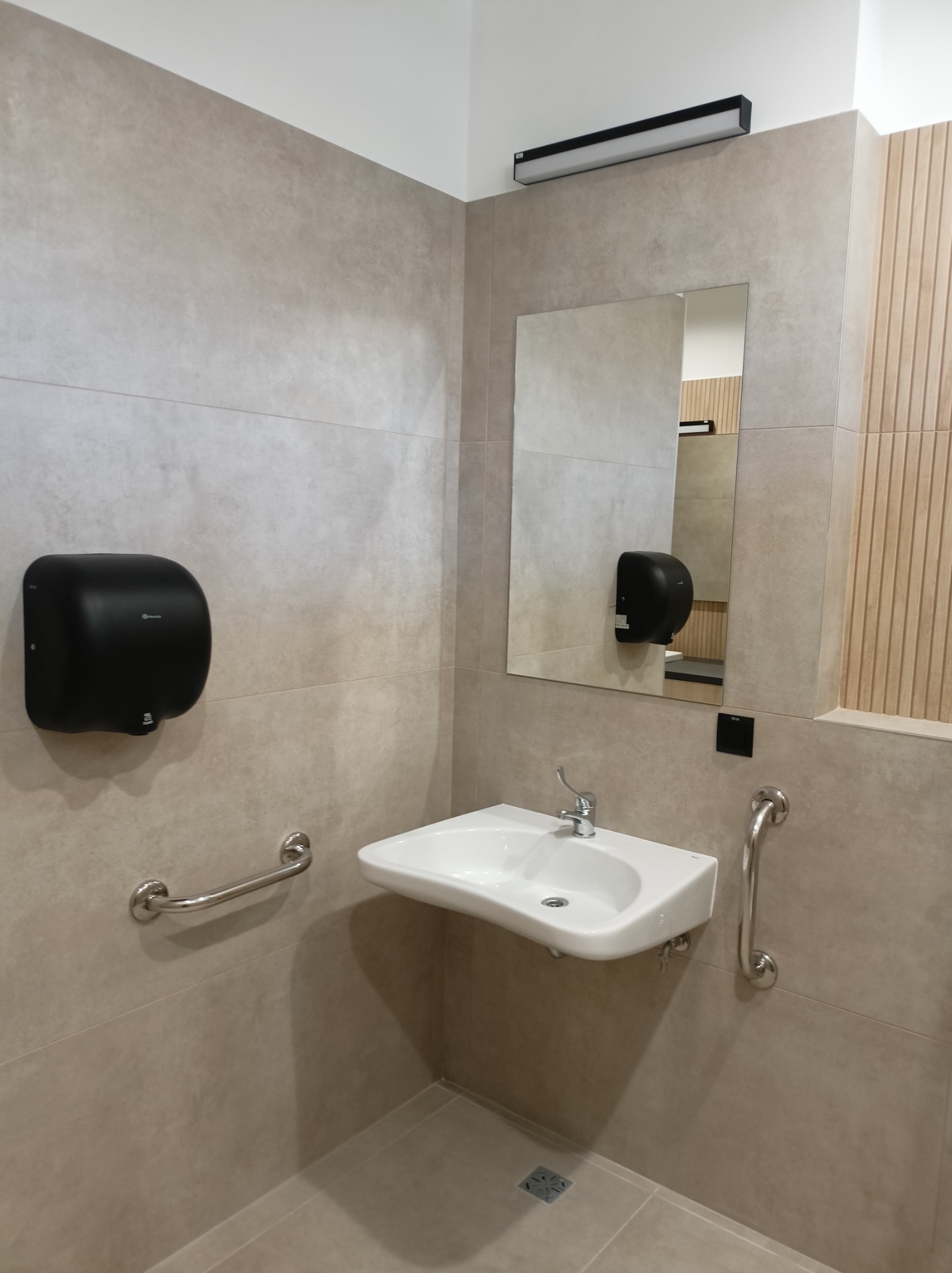 Nowoczesna łazienka publiczna z umywalką, lustrem i uchwytami dla niepełnosprawnych. Beżowe płytki i dyskretne oświetlenie tworzą minimalistyczny design.