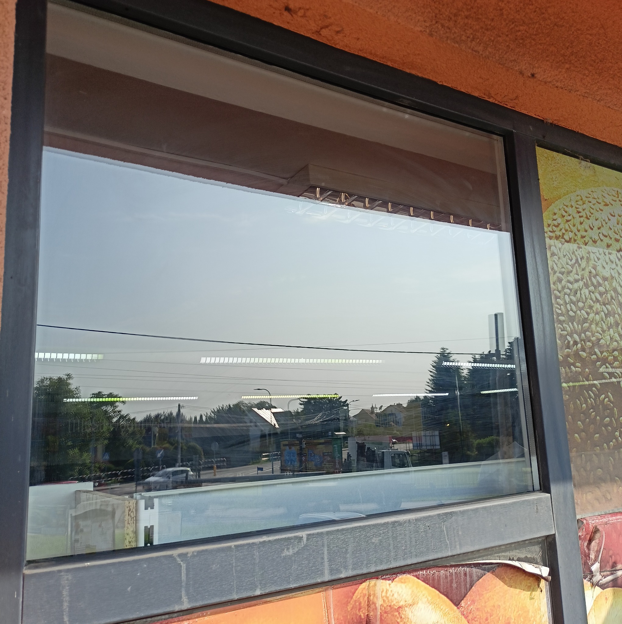 Szklana szyba okienna z widokiem na ulicę, odbijająca otoczenie. Widoczne drzewa i budynki. Szara rama okienna z fragmentem reklamy po prawej.