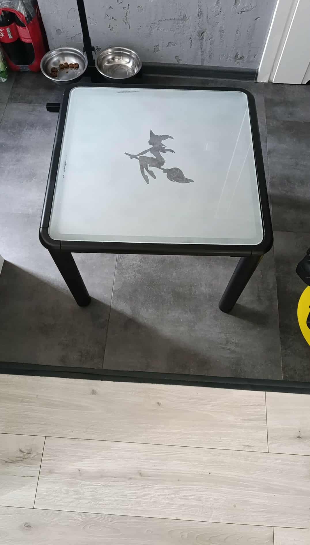 Kwadratowy stół z blatem ze szkła, na którym widnieje motyw czarownicy na miotle. Czarna rama i nogi stołu. W tle miski dla zwierząt i podłoga.