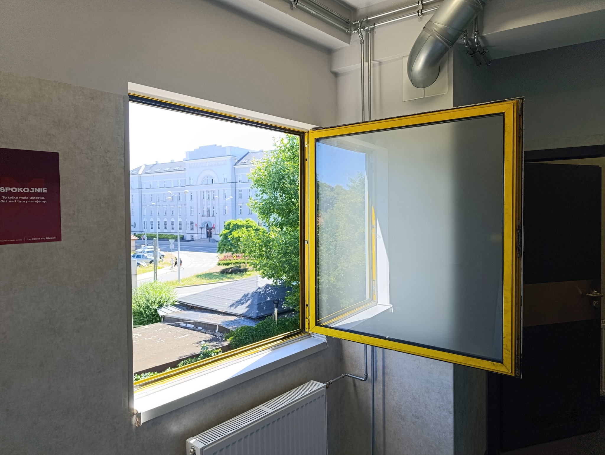 Otworzone okno z żółtą ramą, widok na budynek miejski i zieleń. Wnętrze z grzejnikiem i plakatem. Elementy instalacji na suficie.