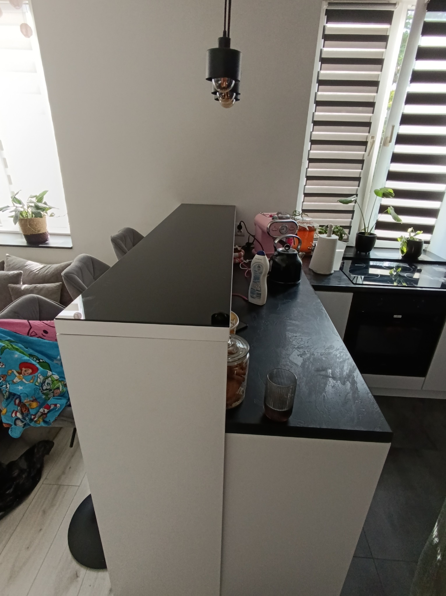 Nowoczesna kuchnia z czarnym blatem i białymi szafkami, widok na okno z roletami i rośliny doniczkowe. Minimalistyczny design, oświetlenie punktowe.