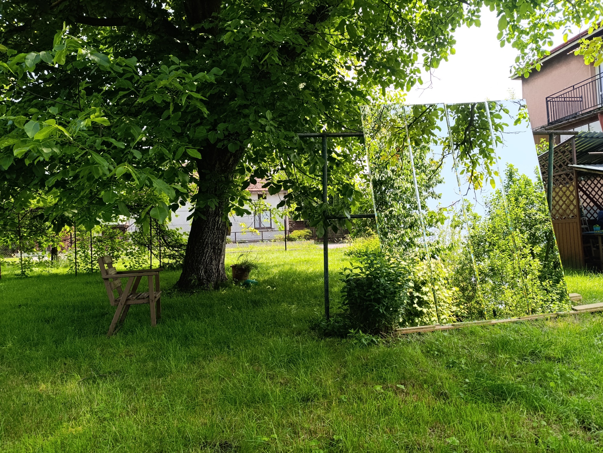 Duże lustro ustawione w ogrodzie, odbija drzewa i budynki. Widoczny drewniany stelaż podtrzymujący taflę, trawa i zieleń wokół. Nietypowe użycie lustra.