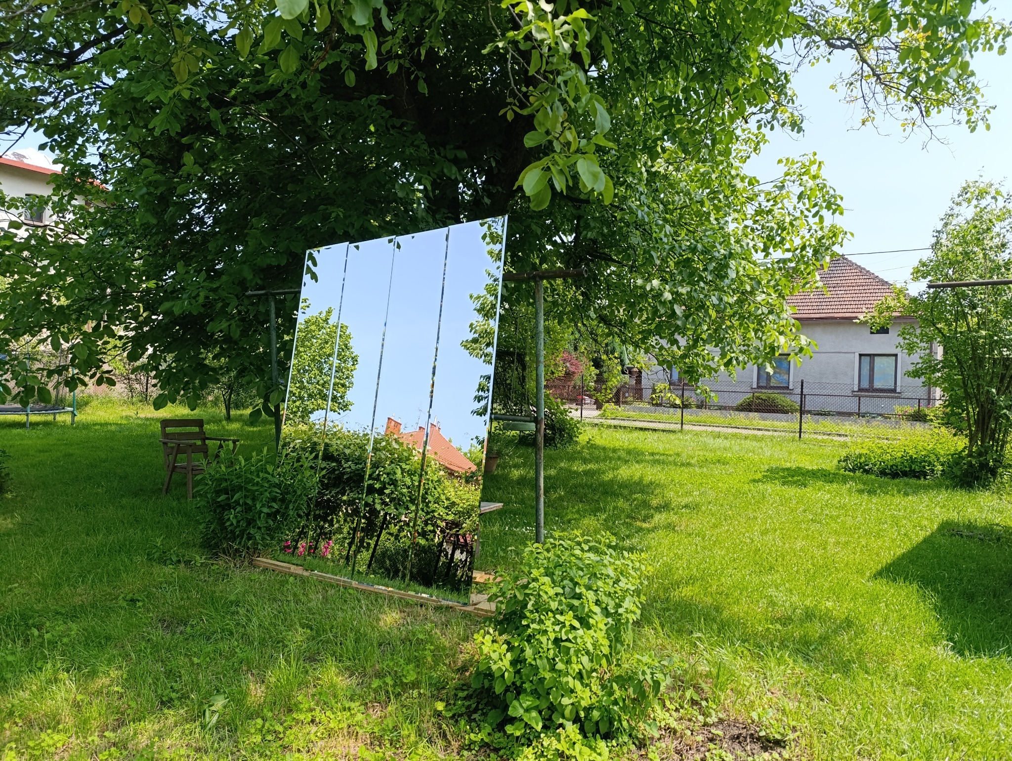 Szklana tafla ustawiona w ogrodzie odbija niebo i zieleń, tworząc iluzję przestrzeni. Widoczne łączenia tafli podkreślają jej konstrukcję. W tle dom i drzewa.