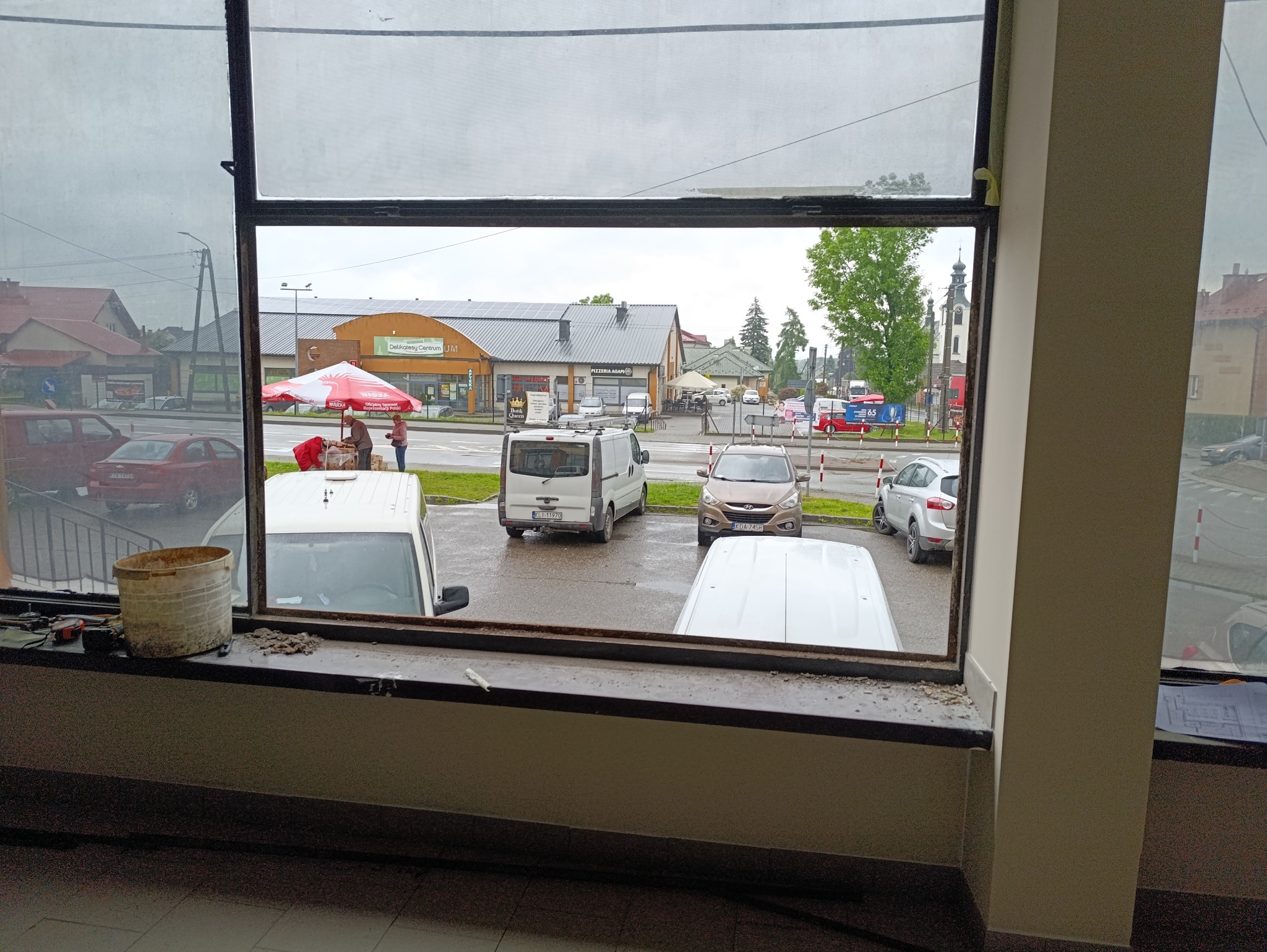 Widok z okna w trakcie montażu, z narzędziami i wiadrem na parapecie. Na zewnątrz budynki i zaparkowane samochody.