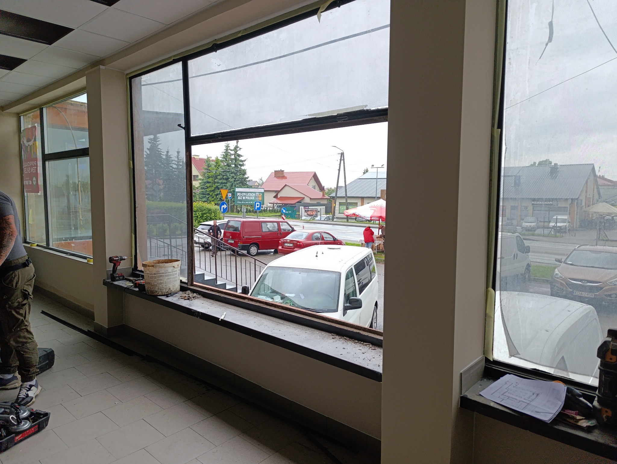 Montaż okna: widok z wnętrza na ulicę przez surowe ramy okienne. Narzędzia i plany na parapecie, sylwetka osoby po lewej. Etap prac szklarskich.