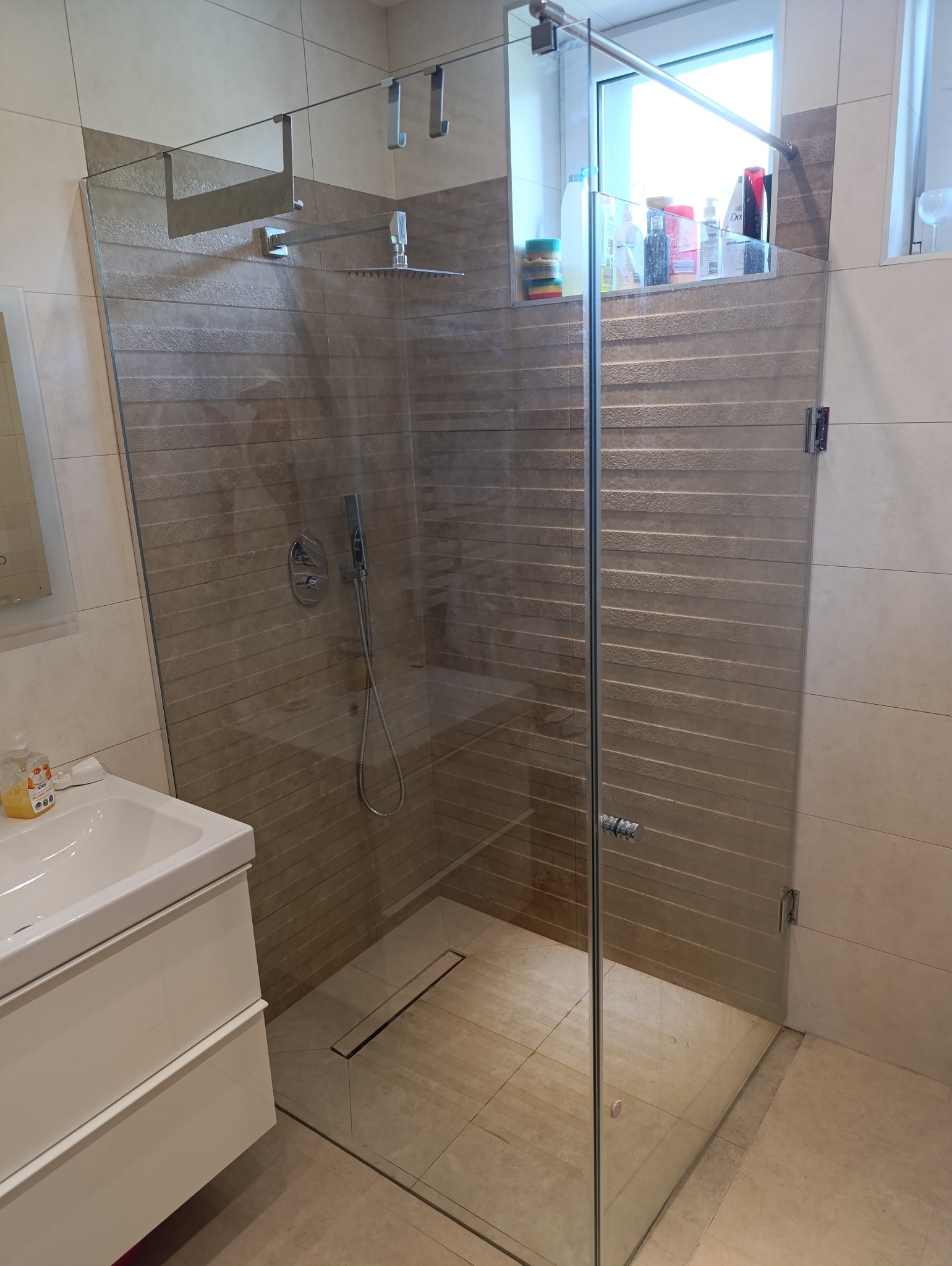Kabina prysznicowa ze szkła hartowanego z deszczownicą, zamontowana w łazience z beżowymi płytkami. Widoczny odpływ liniowy i umywalka z szafką.