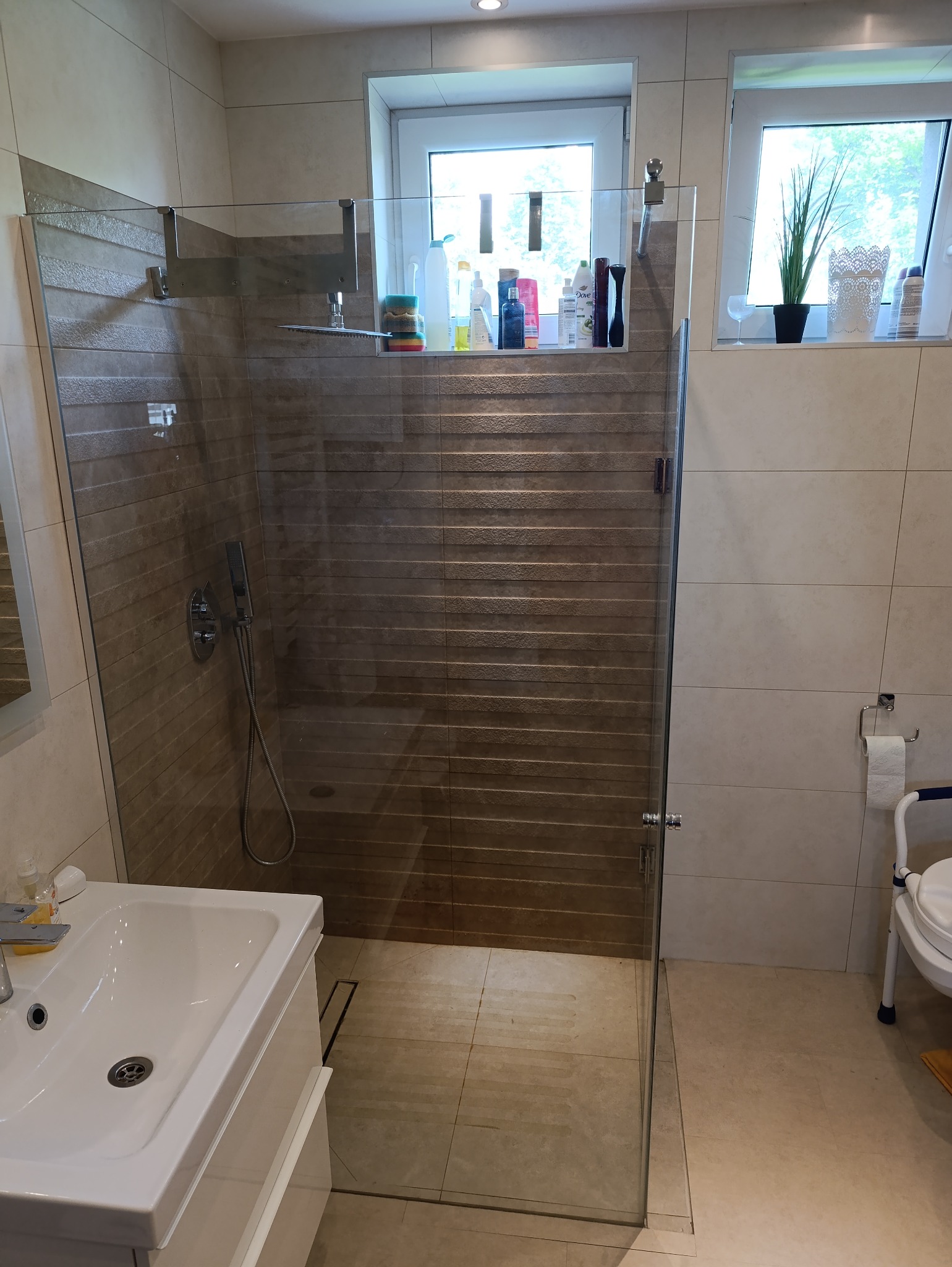 Nowoczesna kabina prysznicowa ze szkła hartowanego, z widoczną armaturą i odpływem liniowym w podłodze. Jasne płytki i okno z rośliną doniczkową dopełniają aranżację łazienki.