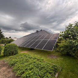 Caldo - Czarny panel słoneczny na zielonym trawniku, na tle domu z ciemnym dachem i pochmurnego nieba. Instalacja w ogrodzie.