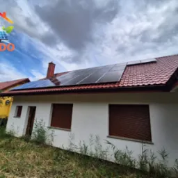 Dom jednorodzinny z instalacją paneli fotowoltaicznych na czerwonym dachu. Widoczne brązowe rolety okienne i logo firmy w lewym górnym rogu.