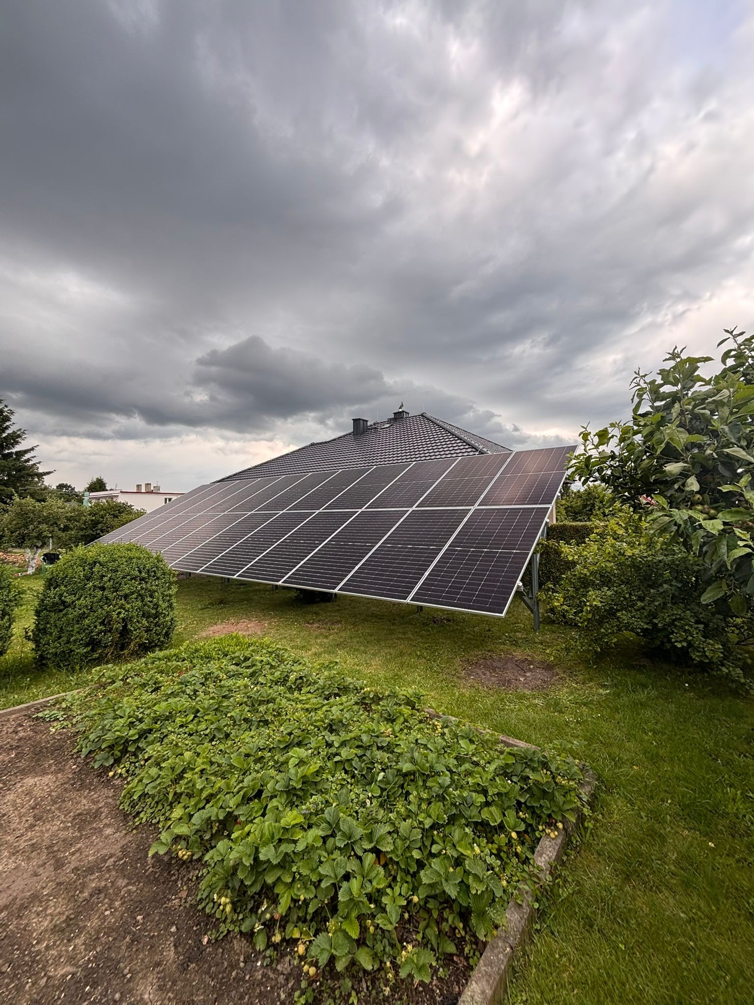Czarny panel słoneczny na zielonym trawniku, na tle domu z ciemnym dachem i pochmurnego nieba. Instalacja w ogrodzie.