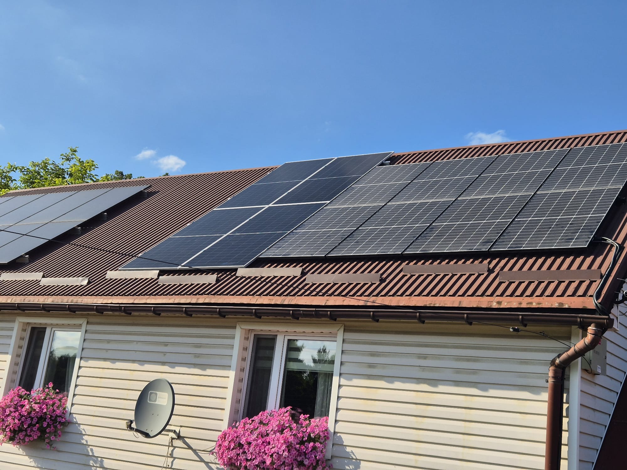 Panele na dachu z brązowej blachy trapezowej, biały dom z kwiatami w oknach i anteną satelitarną, błękitne niebo.