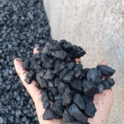 Dłoń trzymająca bryły czarnego węgla na tle betonowej ściany i hałdy węgla, z widoczną fakturą i detalami. Prezentacja produktu.