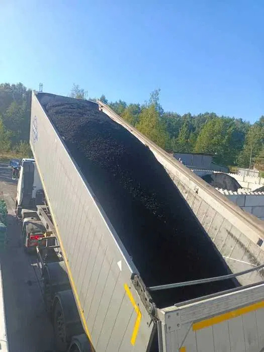 Ciężarówka wyładowuje czarny materiał sypki, prawdopodobnie węgiel, na tle zieleni i błękitnego nieba, z widocznymi elementami konstrukcyjnymi i pojazdami.