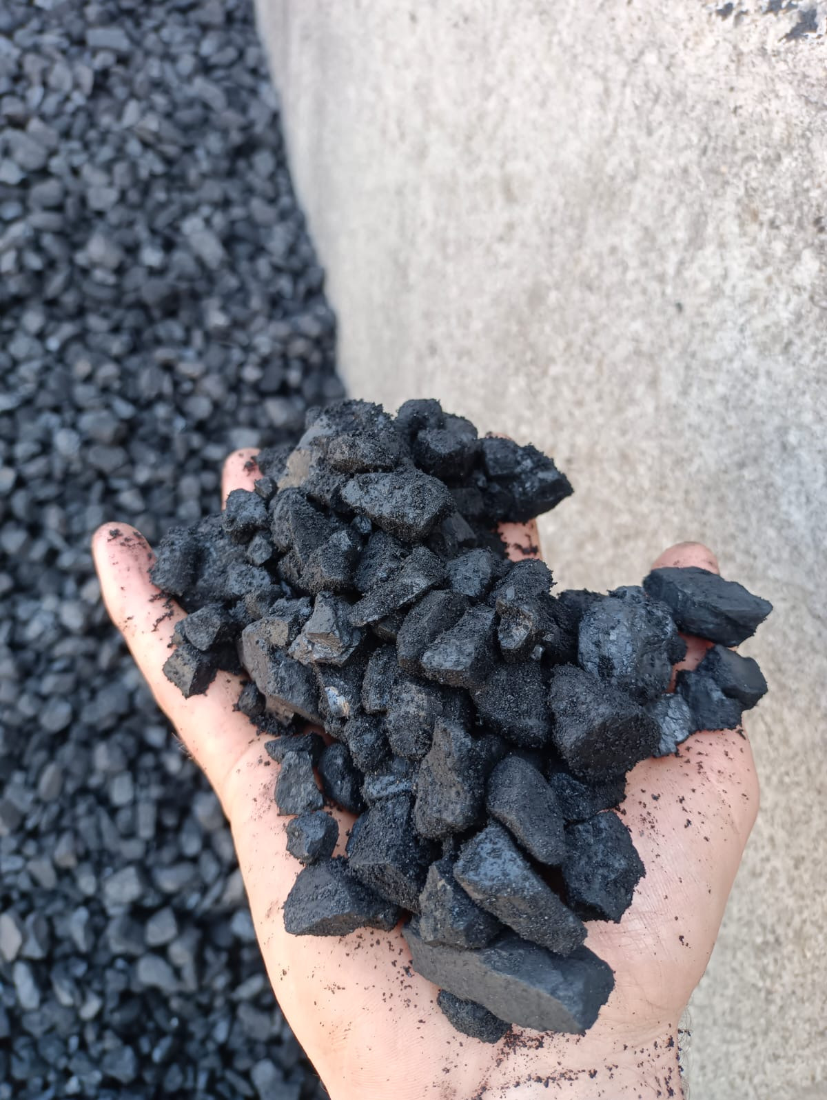 Dłoń trzymająca bryły czarnego węgla na tle betonowej ściany i hałdy węgla, z widoczną fakturą i detalami. Prezentacja produktu.