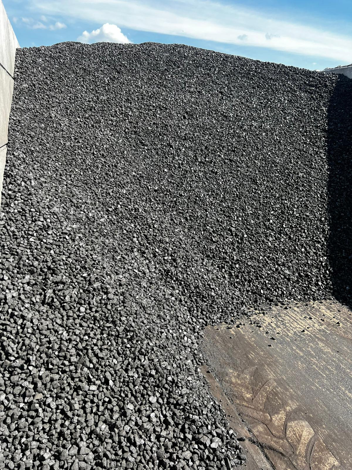 Góra czarnego węgla kamiennego pod błękitnym niebem, widoczna tekstura i pojedyncze bryłki, zarys betonowej ściany po lewej stronie.