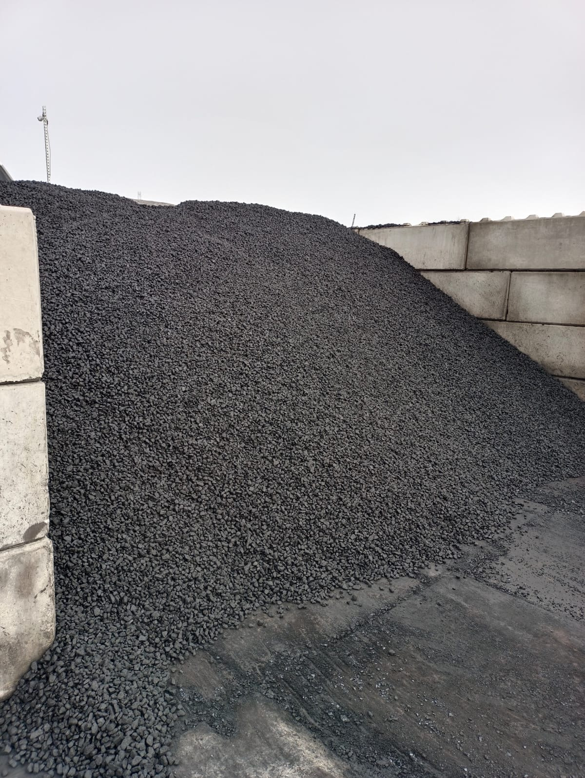 Góra antracytu na składzie, widoczna tekstura i bryły węgla. Tło stanowią betonowe bloki i pochmurne niebo. Ujęcie z dołu podkreśla skalę.