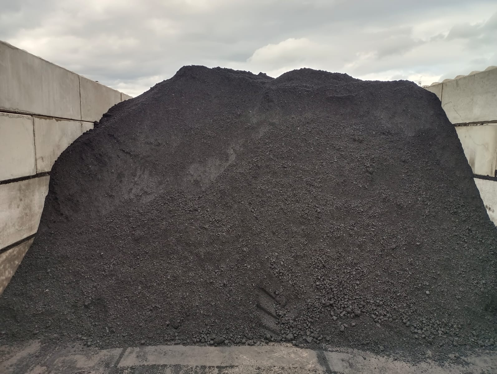 Góra węgla sortowanego na składzie, widoczna tekstura i frakcja, w tle betonowe ściany, pochmurne niebo. Ślady opon w dolnej części hałdy.