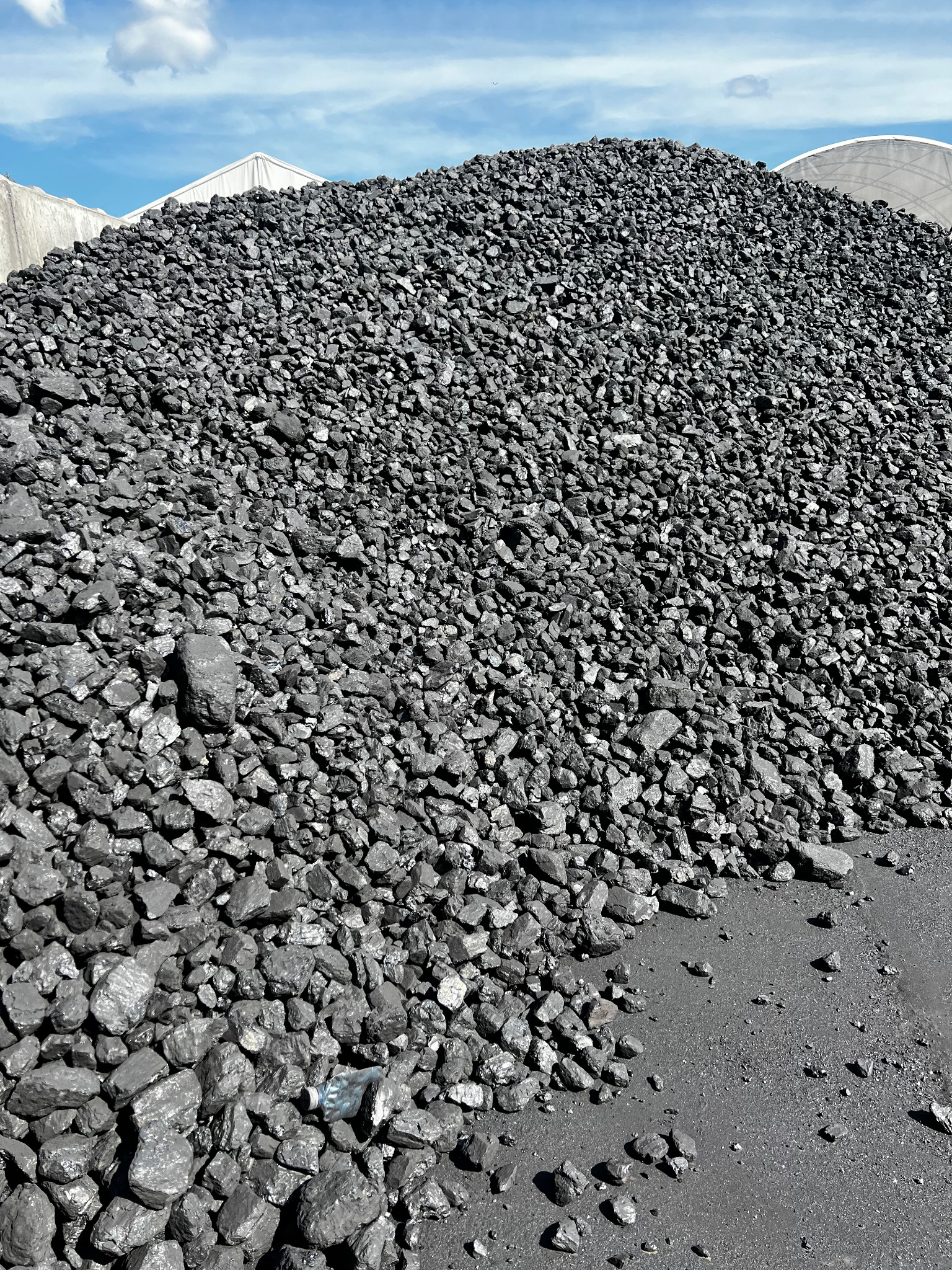 Góra antracytu pod błękitnym niebem z chmurami, widoczna część hali magazynowej w tle. W dolnej części zdjęcia drobny gruz węglowy i plastikowa butelka.