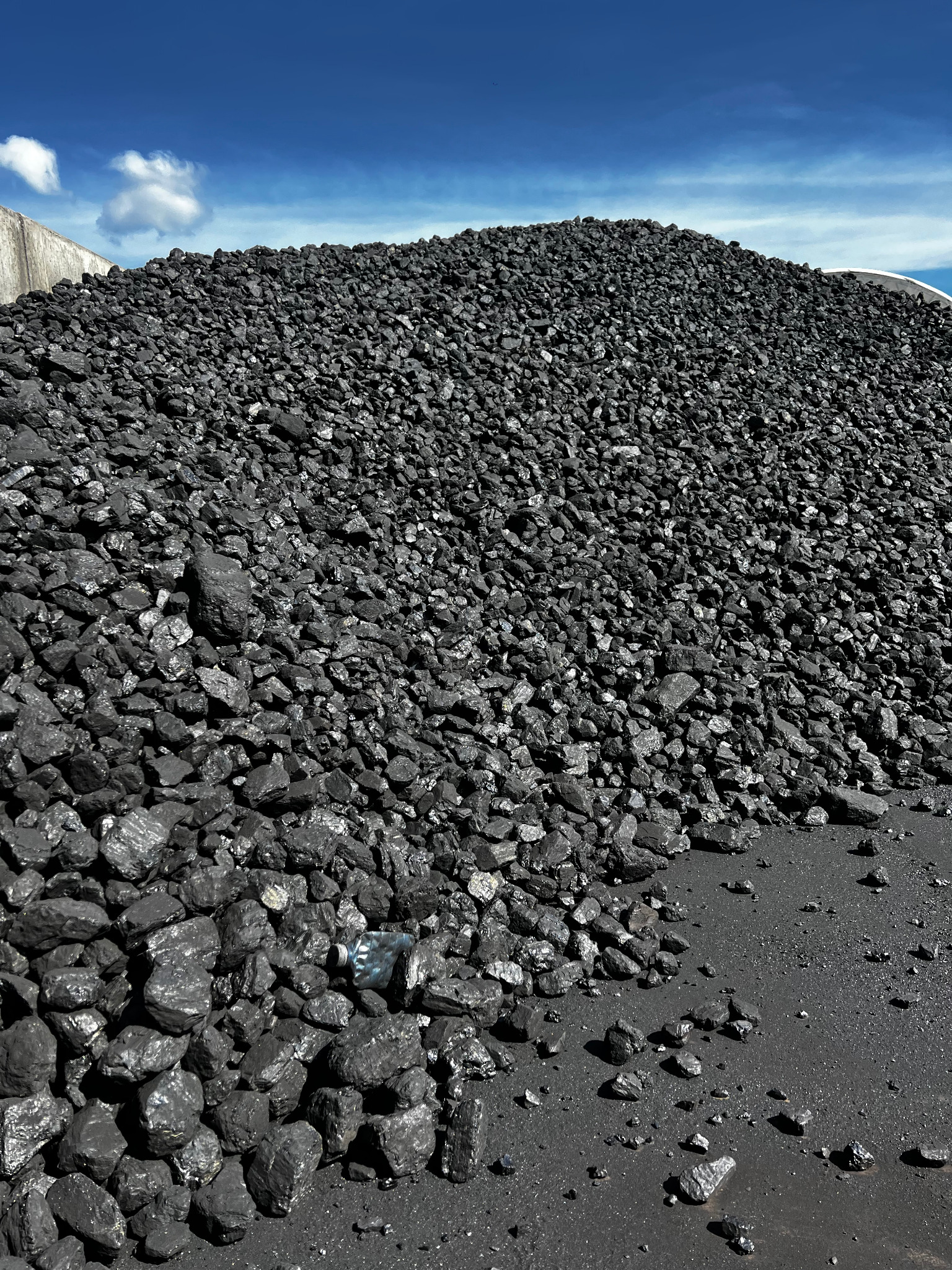Góra czarnego węgla kamiennego pod błękitnym niebem, z widoczną plastikową butelką u podstawy hałdy. Widok z dołu, podkreślający teksturę i objętość.
