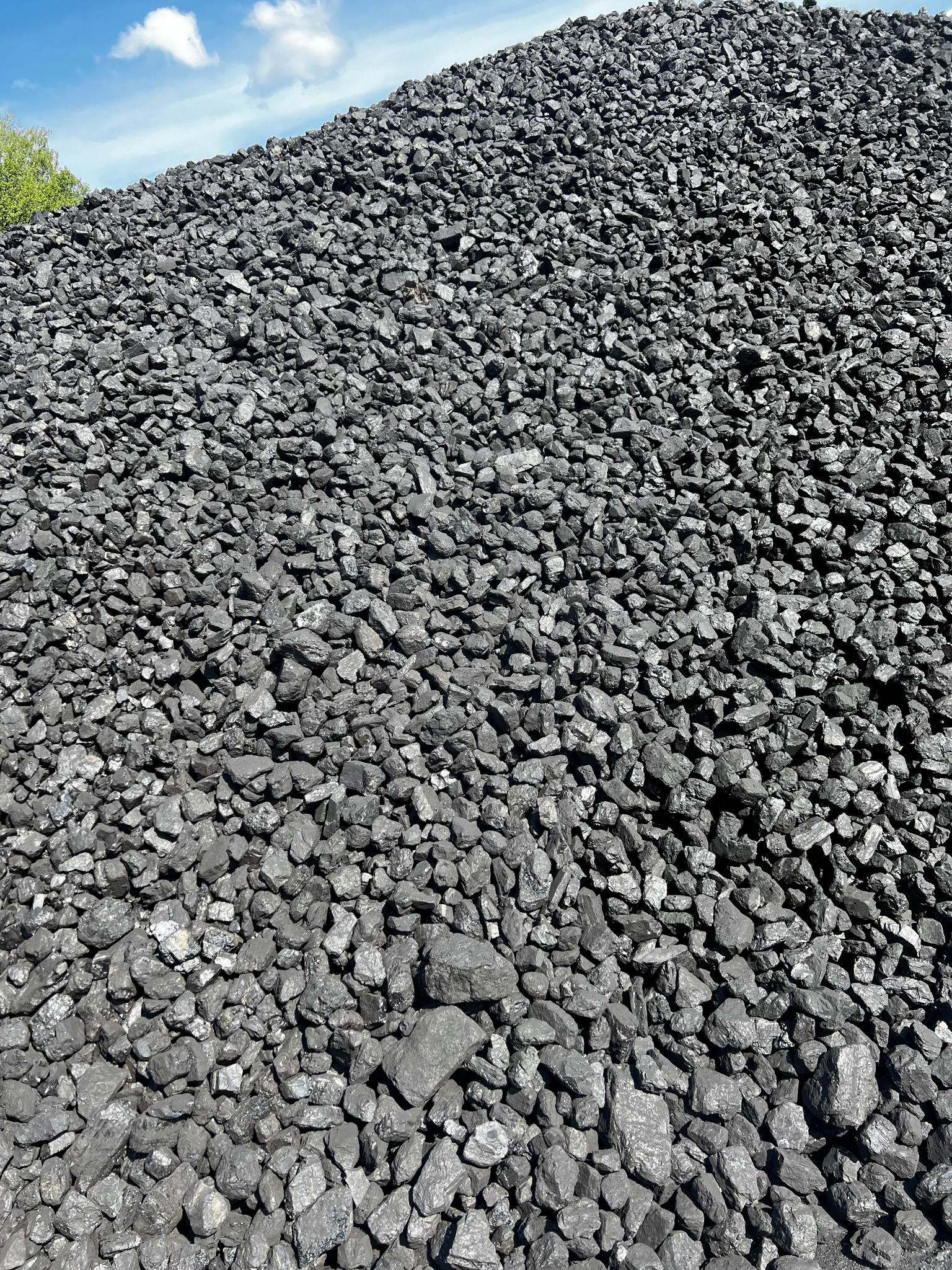 Góra czarnego węgla kamiennego pod błękitnym niebem z chmurami, widoczna zieleń drzew po lewej stronie, z bliska, w pełnym słońcu.