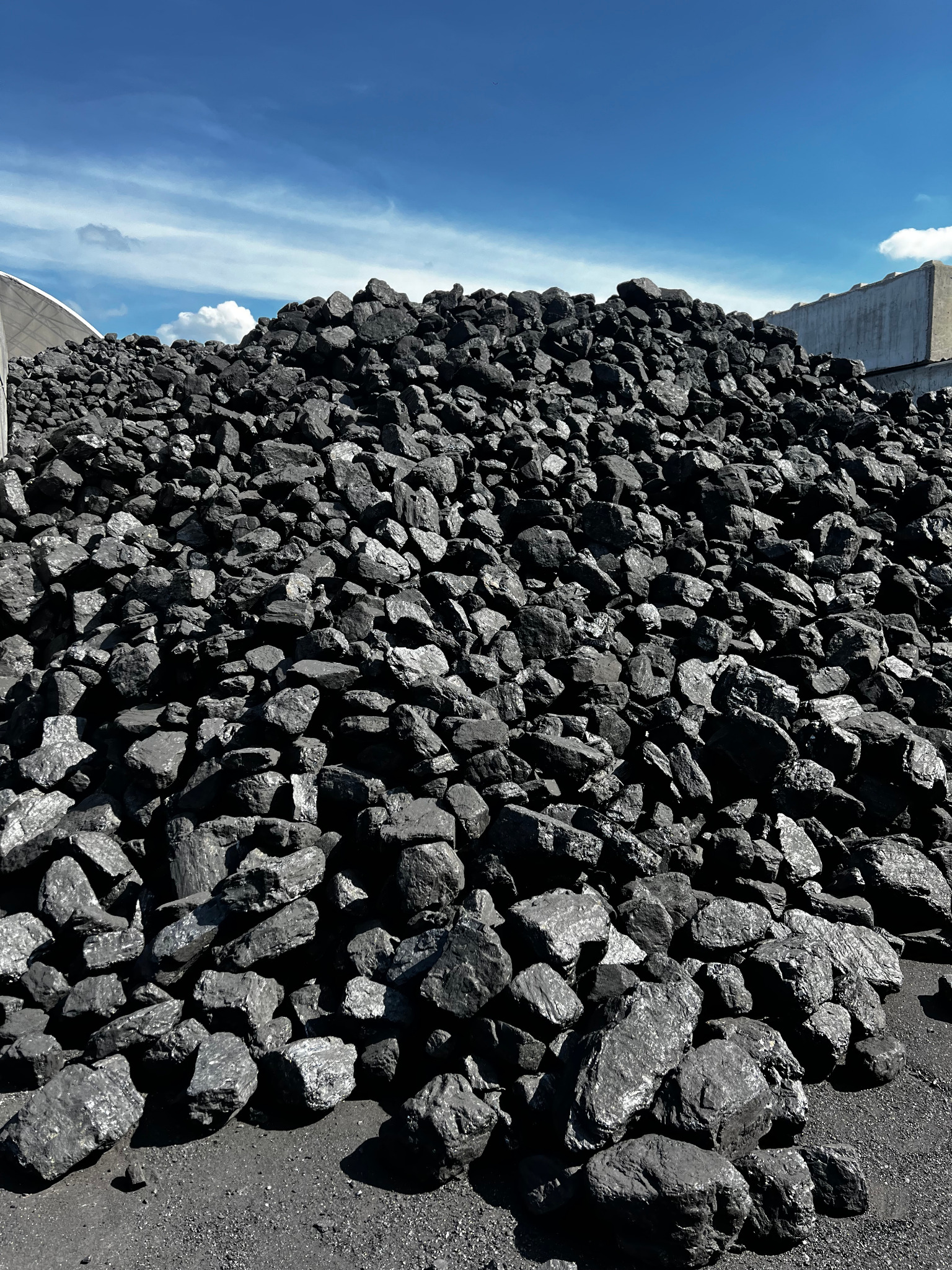 Góra czarnego węgla kamiennego pod błękitnym niebem. Widoczna betonowa konstrukcja w tle. Ujęcie z dołu, podkreślające objętość materiału.
